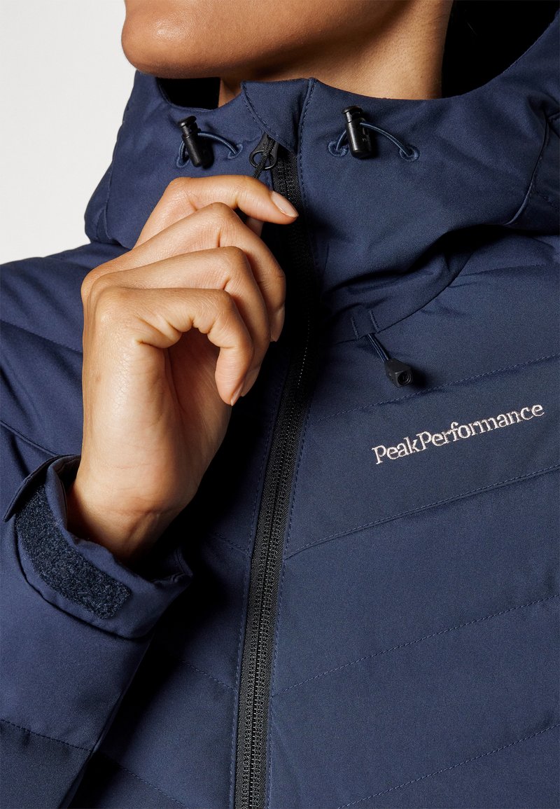 Dunkelblaue isolierte Jacke mit hohem Kragen, ausgestattet mit einem Frontreißverschluss, verstellbaren Kordelzügen und Klettverschlussärmeln. Logo "PeakPerformance" sichtbar.