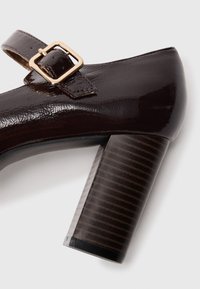 Brown patent leather heeled shoe with a square toe, adjustable strap featuring a gold buckle, and a patterned, chunky heel.