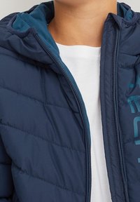 Granatowa puchowa kurtka o pikowanej teksturze, kontrastowe tealowe wnętrze, kaptur i zamek błyskawiczny. Posiada nadrukowany wzór z przodu.