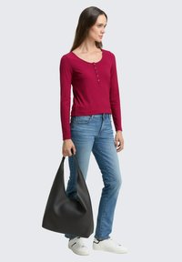 Burgunderfarbener gerippter Langarmtop mit rundem Halsausschnitt und Knopfdetails, kombiniert mit blauen Jeans und einer großen schwarzen Hobo-Handtasche, die in einer Hand gehalten wird.
