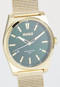 Reloj en tono dorado con una esfera verde, correa de malla, marcadores de hora en relieve y una característica de resistencia al agua etiquetada como "5 BAR". Nombre de la marca "HUGO" visible.
