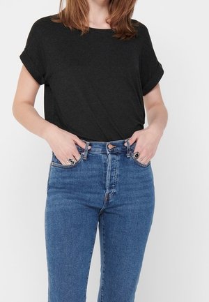 Vrouw draagt een zwart T-shirt met korte mouwen ingestopt in een high-waisted blauwe spijkerbroek, met handen die rusten bij de zakken van de spijkerbroek en ringen aan de vingers.