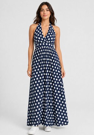 Donna con abito lungo senza maniche blu navy a pois bianchi abbinato a scarpe da ginnastica bianche, in piedi davanti a uno sfondo semplice.