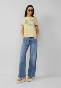 Geel T-shirt met korte mouwen en "Aloha" grafiek, gecombineerd met lichtblauwe wijde spijkerbroek en crèmewitte platform sandalen.
