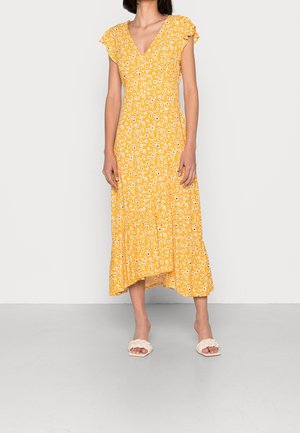 Femme portant une robe midi jaune à fleurs avec des manches courtes volantées et des sandales slides blanches tressées, debout devant un fond uni.