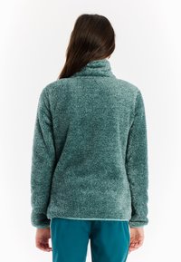 Žena u tirkiznoj plišanoj jakni i odgovarajućim hlačama, prikazana s leđa na bijeloj pozadini.