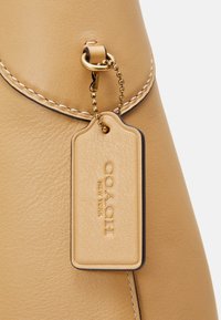 Sac Coach en cuir tan avec des bords cousus et une étiquette assortie en cuir embossée de "COACH NEW YORK" suspendue à une chaîne dorée.