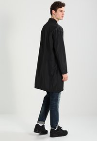 Sort letvægtsjakke med langt lige design, minimal krave, højde til hoften, båret over blå jeans og sorte sneakers, glat tekstur.