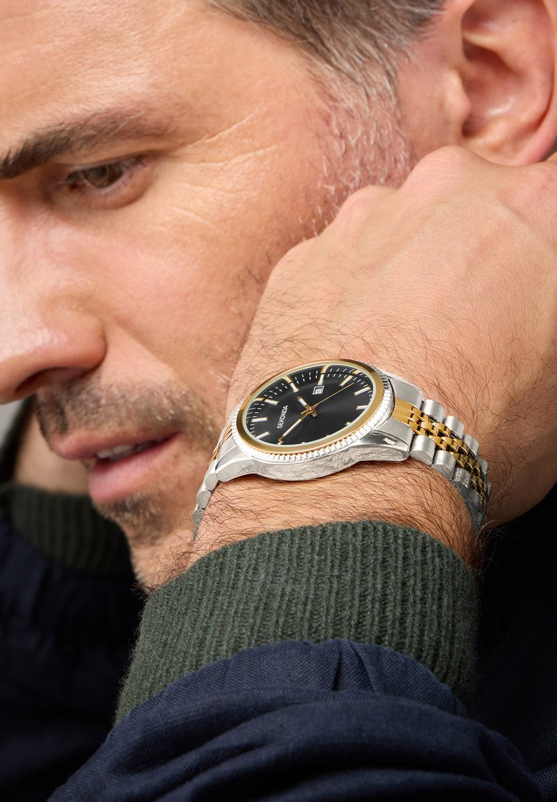 Silver and gold two-tone wristwatch with a black dial, date display, and a textured bezel, worn on a man's wrist.