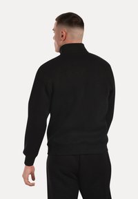 Hombre con pelo corto que lleva un sudadera negra de cuello alto y pantalones negros, visto desde atrás contra un fondo simple.