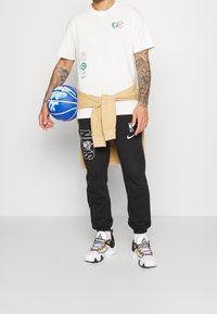 Vit t-shirt med logotyper, svarta joggers med "Nets"-tryck, beiget tröja knuten i midjan, och flerfärgade basketsko-accenter.