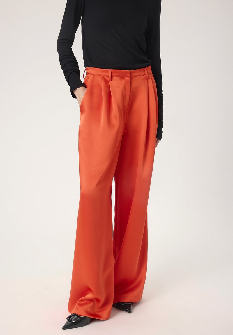 Pantalons large en satin orange avec des plis et des poches latérales, associés à un haut noir à manches longues, portés avec des chaussures noires à bouts pointus.
