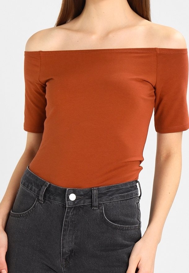 Haut orange rouille épaules dénudées avec manches courtes associé à un jean en denim noir taille haute porté par une personne sur un fond uni.