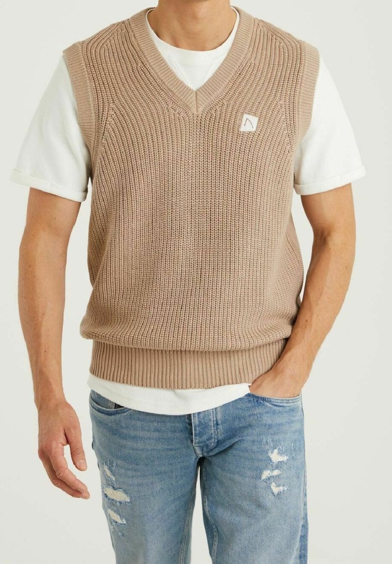 Light brown knitted vest with a v-neck design, ribbed texture, and a small logo patch. Worn over a white t-shirt and blue distressed jeans.