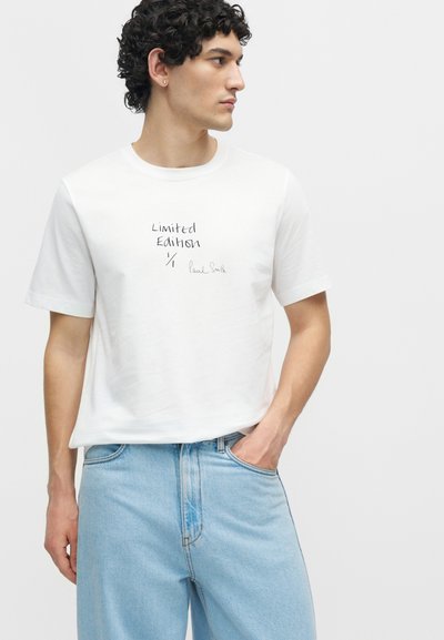 Νέος άνδρας με σγουρά μαλλιά που φοράει λευκό μπλουζάκι με το κείμενο "Limited Edition" και ανοιχτό μπλε τζιν, κοιτάζει προς τα δεξιά με το ένα χέρι στην τσέπη.