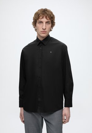 Junger Mann mit lockigem Haar, der ein schwarzes Hemd mit Knöpfen und graue Hosen trägt, steht vor einem schlichten weißen Hintergrund.