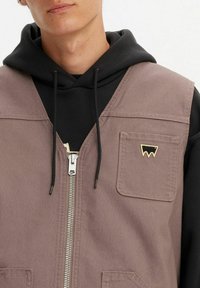 Gilet in tela marrone con una cerniera argentata, tasca sul petto con un emblema dorato, indossato sopra un felpa nera con cappuccio e lacci.