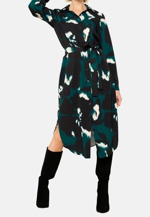 Frau trägt ein langärmliges, türkis-schwarzes gemustertes Hemdkleid mit Stoffgürtel, kombiniert mit schwarzen kniehohen Stiefeln, posiert mit einem erhobenen Arm.