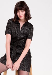 Young woman with short brown hair sitting on wooden stool, wearing short-sleeve black textured dress with front zipper and tie waist.