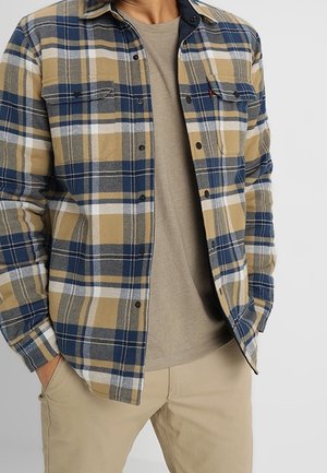 Mężczyzna w niebiesko-beżowo-biało-czarnej kratowanej koszuli zapinanej na guziki, na beżowym t-shircie i beżowych spodniach, ręce w kieszeniach.