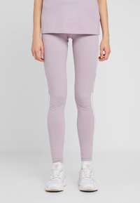 Světle fialové legíny vyrobené z elastické tkaniny s hladkou strukturou, s bílými bočními pruhy a logem poblíž kotníku.