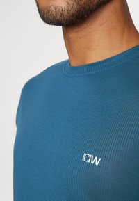 Blå teksturert sportsskjorte med rund hals og kortermet, med en hvit logo "ICIW" tydelig plassert på brystet.