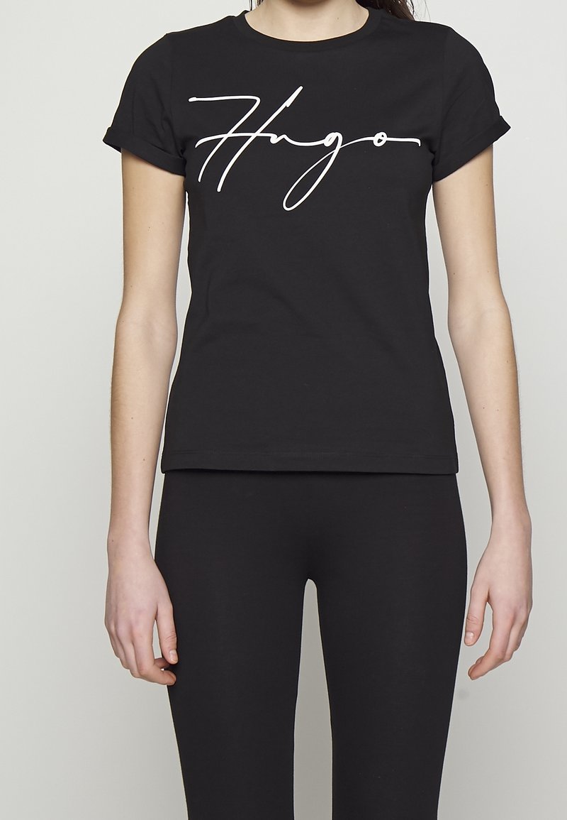 T-shirt en coton noir à manches courtes roulées, avec un logo "Hugo" en cursive blanche sur le devant, design ajusté, texture lisse.