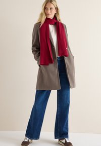 Bruine wollen overjas, witte shirt, blauwe wijde jeans en een geribde rode sjaal. Sneakers hebben crèmekleurige en bruine accenten.