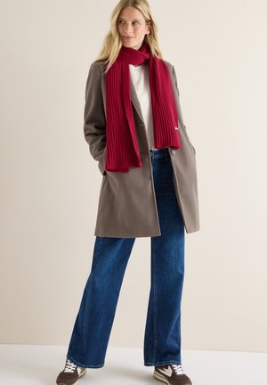 Brun ullblandad ytterrock, vit skjorta, blå vida jeans och en ribbad röd scarf. Sneakers har krämfärgade och bruna detaljer.