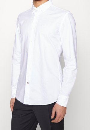 Muž v dlouhém rukávu bílé košile se zapínáním na knoflíky a špičatým límečkem a černých kalhotách, stojící před jednoduchým pozadím.