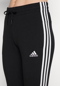 Černé tepláky z měkké tkaniny s elastickým páskem na stažení šňůrkou, se třemi bílými pruhy po stranách a vyšívaným logem.