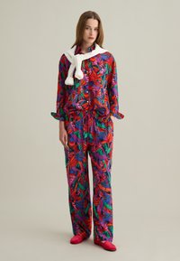 Conjunto de dos piezas que incluye una camisa de manga larga y pantalones. Estampado tropical brillante en rojo, verde y púrpura; suéter blanco atado alrededor del cuello.