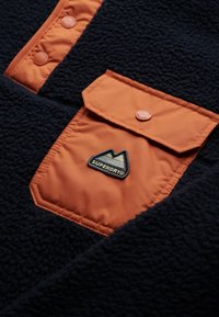 Fleece-Pullover in marineblau mit einer orangenen Nylonbrusttasche, ausgestattet mit einem Druckknopfverschluss und einem schwarzen 'SUPERDRY'-Logo-Patch.