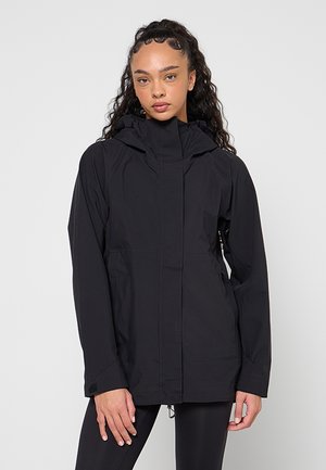 Femme aux longs cheveux bouclés portant une veste à capuche noire et des leggings noirs, debout devant un fond clair uni.