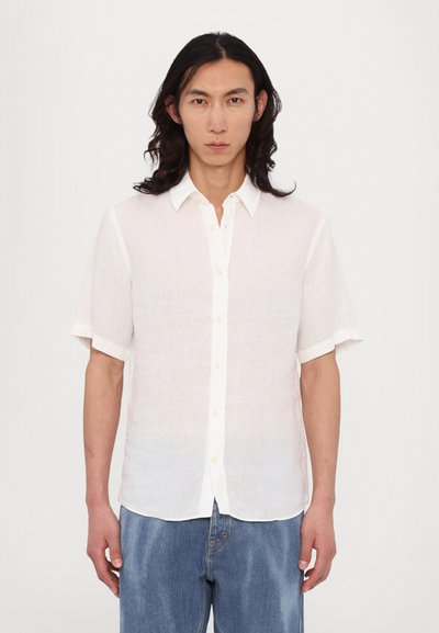 Chemise en lin blanche à manches courtes avec un devant à boutons, présentant un tissu texturé et des accents de boutons clairs contrastants. Associée à un jean en denim bleu.