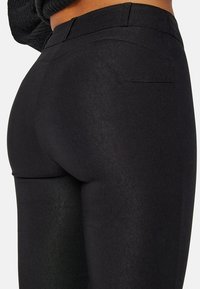 Zwarte aansluitende broek met een gladde textuur, hoge tailleband en een achterzak. De stof heeft een subtiele glans, wat het gestroomlijnde ontwerp accentueert.