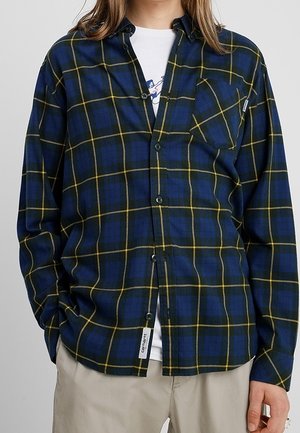 Chemise en flanelle à carreaux marine avec des lignes jaunes, manches longues, fermeture à boutons sur le devant et poche poitrine. Présente une étiquette tissée sur l'ourlet.