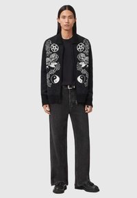 Zwarte gebreide cardigan met witte grafische ontwerpen van rozen, yin-yang symbolen en pentagrammen, gecombineerd met zwarte wijde jeans en laarzen.