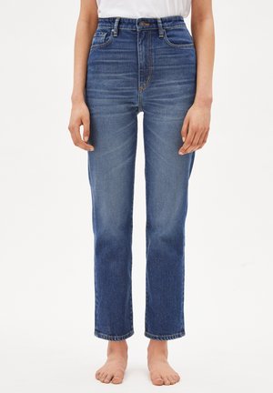 Barfodet person iført blå denimjeans med lige ben og høj talje samt en hvid skjorte, stående mod en ensfarvet hvid baggrund.