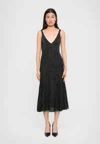 Vestido preto, sem mangas, de decote em V com uma textura brilhante, saia plissada e barra ondulada, combinado com saltos pretos de bico fino.