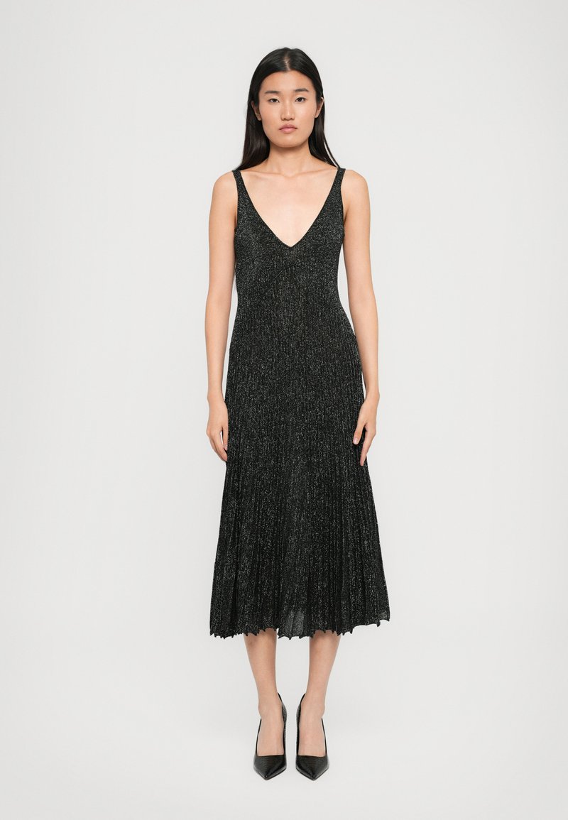 Vestido preto, sem mangas, de decote em V com uma textura brilhante, saia plissada e barra ondulada, combinado com saltos pretos de bico fino.