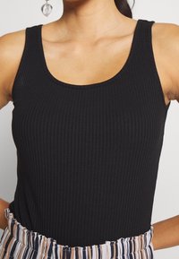 Top nero a costine con scollatura a barchetta e spalline larghe, abbinato a una cintura a righe. Texture liscia e design aderente.
