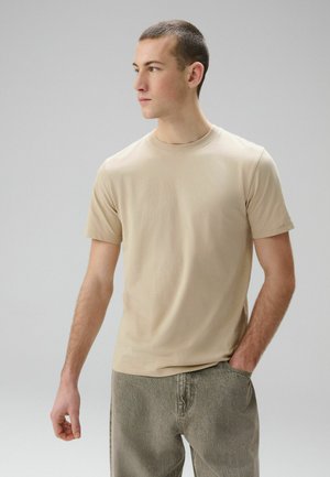 Tânăr purtând un tricou bej cu mâneci scurte și blugi gri, stând cu o mână în buzunar, privind în lateral, pe un fundal simplu.