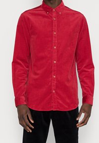 Camisa de corduroy roja con cuello botón, mangas largas y botones dorados a lo largo de la hebilla delantera. Textura notable del tejido acanalado.