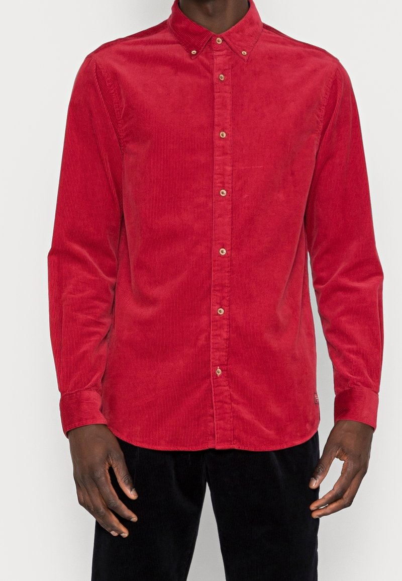 Camisa de corduroy roja con cuello botón, mangas largas y botones dorados a lo largo de la hebilla delantera. Textura notable del tejido acanalado.