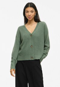 Cardigan en laine vert avec un col en V, cinq boutons marron, des poignets côtelés et une coupe décontractée. Présenté sur un fond neutre.
