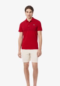 Röd polo med rutmönster över bröstet, med en knäppning med två knappar och en liten logotyp på vänster sida. Matchad med beiga shorts.