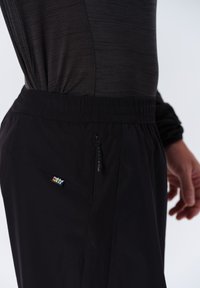 Svarta träningsbyxor har en elastisk midja, ficka med dragkedja och textil med textur och perforeringar för andningsförmåga. Rukka-logotyp synlig.