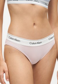 Γυναίκα φορώντας ανοιχτό ροζ εσώρουχα Calvin Klein και ασορτί αθλητικό σουτιέν που δείχνει τη λαστιχένια μέση με το λογότυπο.