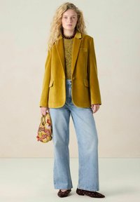 Blazer en velours moutarde avec revers, associé à un haut vert texturé, un jean clair à jambes larges et un sac à main à motifs.